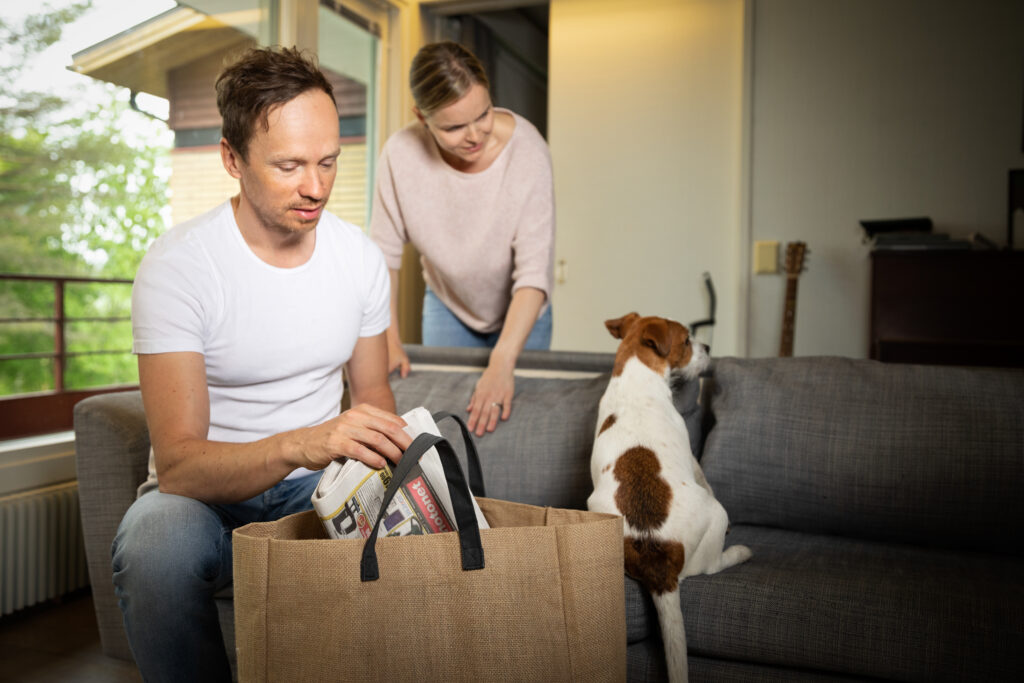 Mies laittaa lehteä kangaskassiin ja nainen katsoo taustalla perheen koiraa