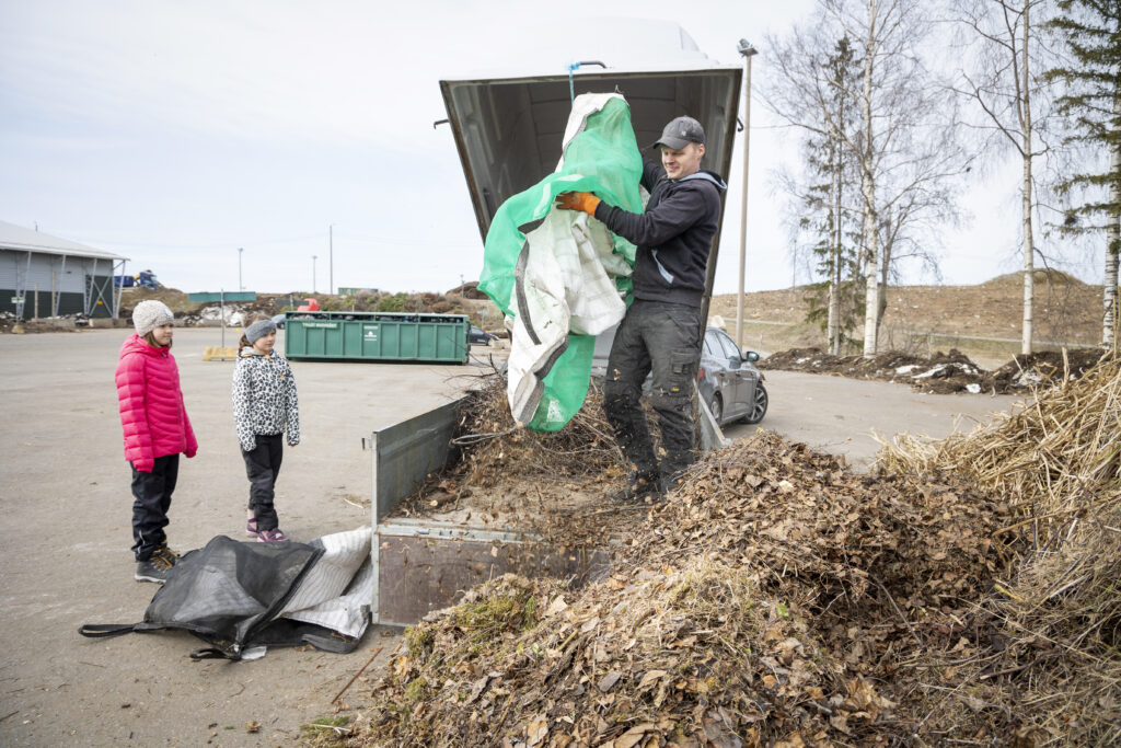 Mies tyhjentää haravointijätettä isosta säkistä, kun lapset katsovat