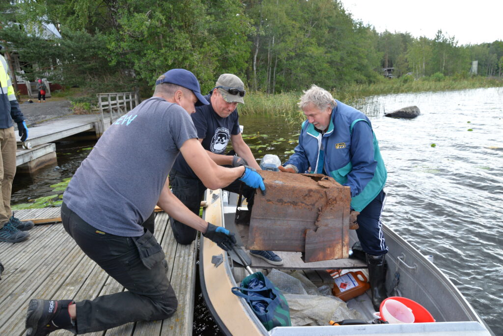 Kolme miestä nostaa vanhaa kiuasta soutuveneestä laiturille