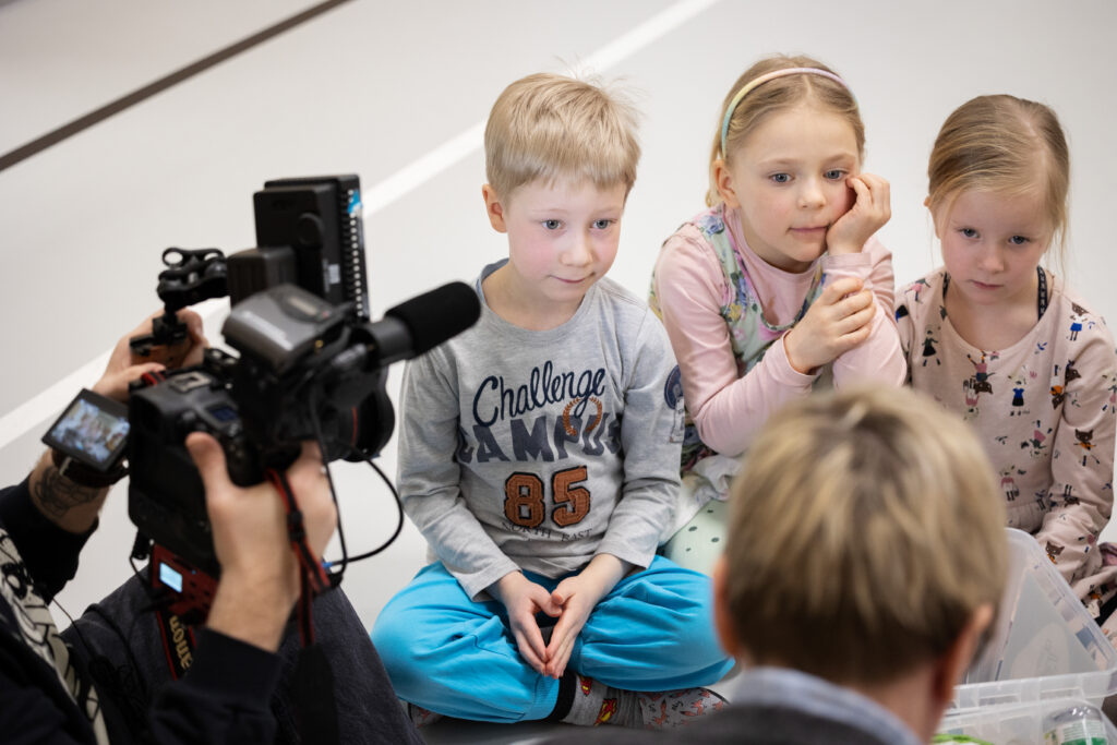 Lapset katsovat toimittajaa, kun videokamera kuvaa
