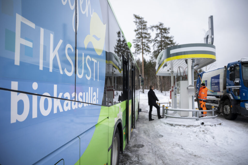 Bussin kyljessä on "Kuljen fiksusti biokaasulla" -tarra, taustalla näkyy kaksi miestä tankkaamassa Seppälän tankkausasemalla