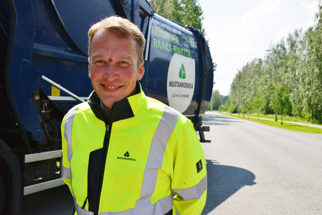 Mies katsoo kameraan hymyillen huomiovaatteissa, taustalla näkyy jäteauto