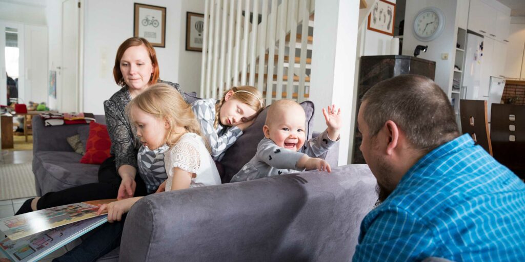 Koko perhe touhuaa sohvalla, isommat tytöt lukevat, pieni vauva ilmeilee isälleen