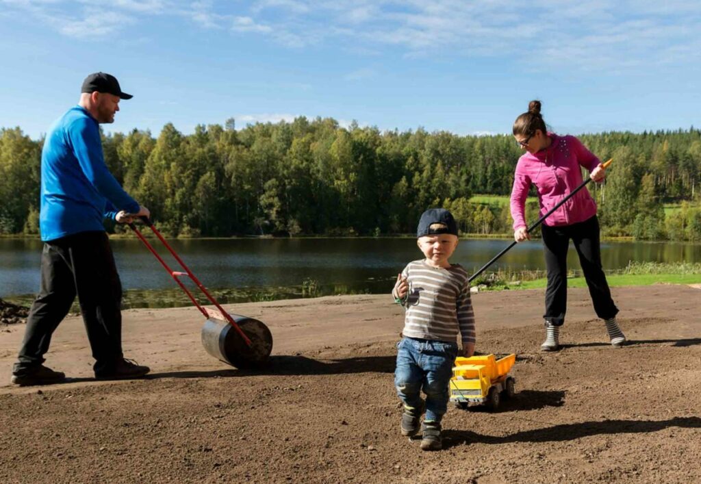 Perheen isä jyrää ja äiti haraa multaa. Perheen poika vetää muovirekkaa narussa.