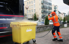 Oranssipukuinen mies pesee painepesurilla keltaista muovijätteiden jäteastiaa.