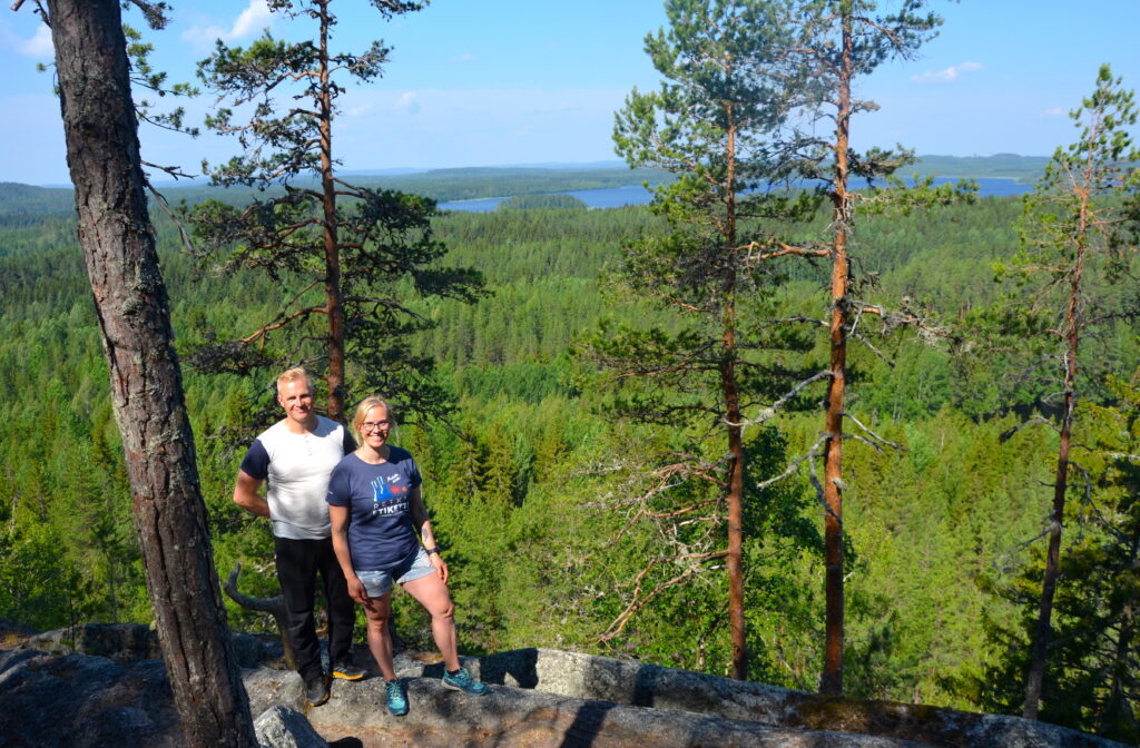 Mies ja nainen seisovat komeissa maisemissa Hyyppäänvuorella, alhaalla näkyy puita ja järveä.