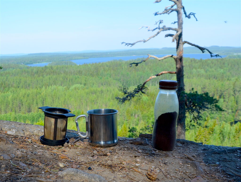 Kahvin kestosuodatin, metallinen kuppi sekä kahvinporoja muovipullossa korkealla mäen päällä.