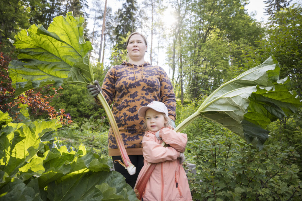 Nainen ja hänen lapsensa pitävät paksuja raparpereja kädessään, taustalla näkyy vihreää puutarhaa.