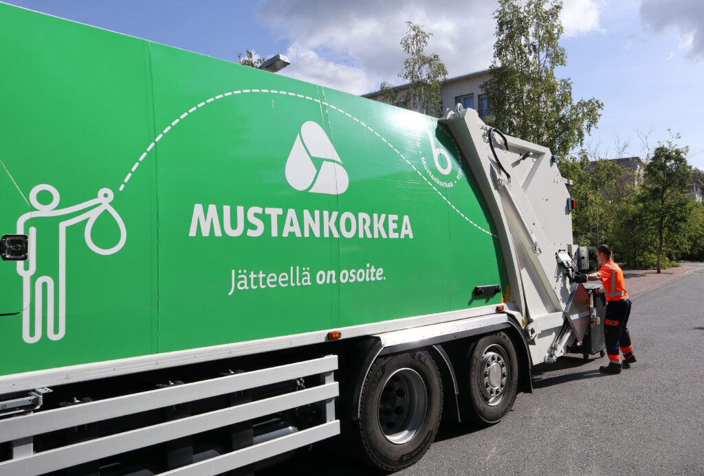 Huomiovaatetukseen pukeutunut mies tyhjentää jäteastiaa, etualalla näkyy vihreä jäteauto.