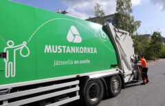 Huomiovaatetukseen pukeutunut mies tyhjentää jäteastiaa, etualalla näkyy vihreä jäteauto.