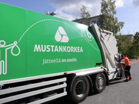 Huomiovaatetukseen pukeutunut mies tyhjentää jäteastiaa, etualalla näkyy vihreä jäteauto.