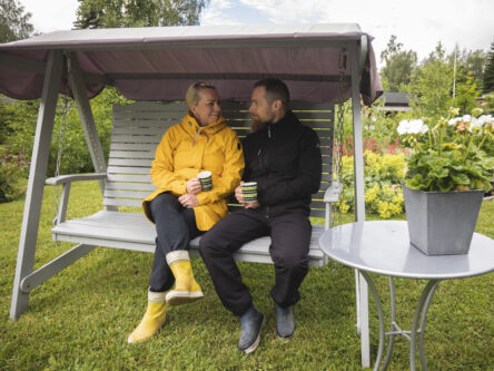Nainen keltaisessa takissa ja mies mustassa takissa istuvat pihakeinussa, taustalla näkyy perennepenkki.
