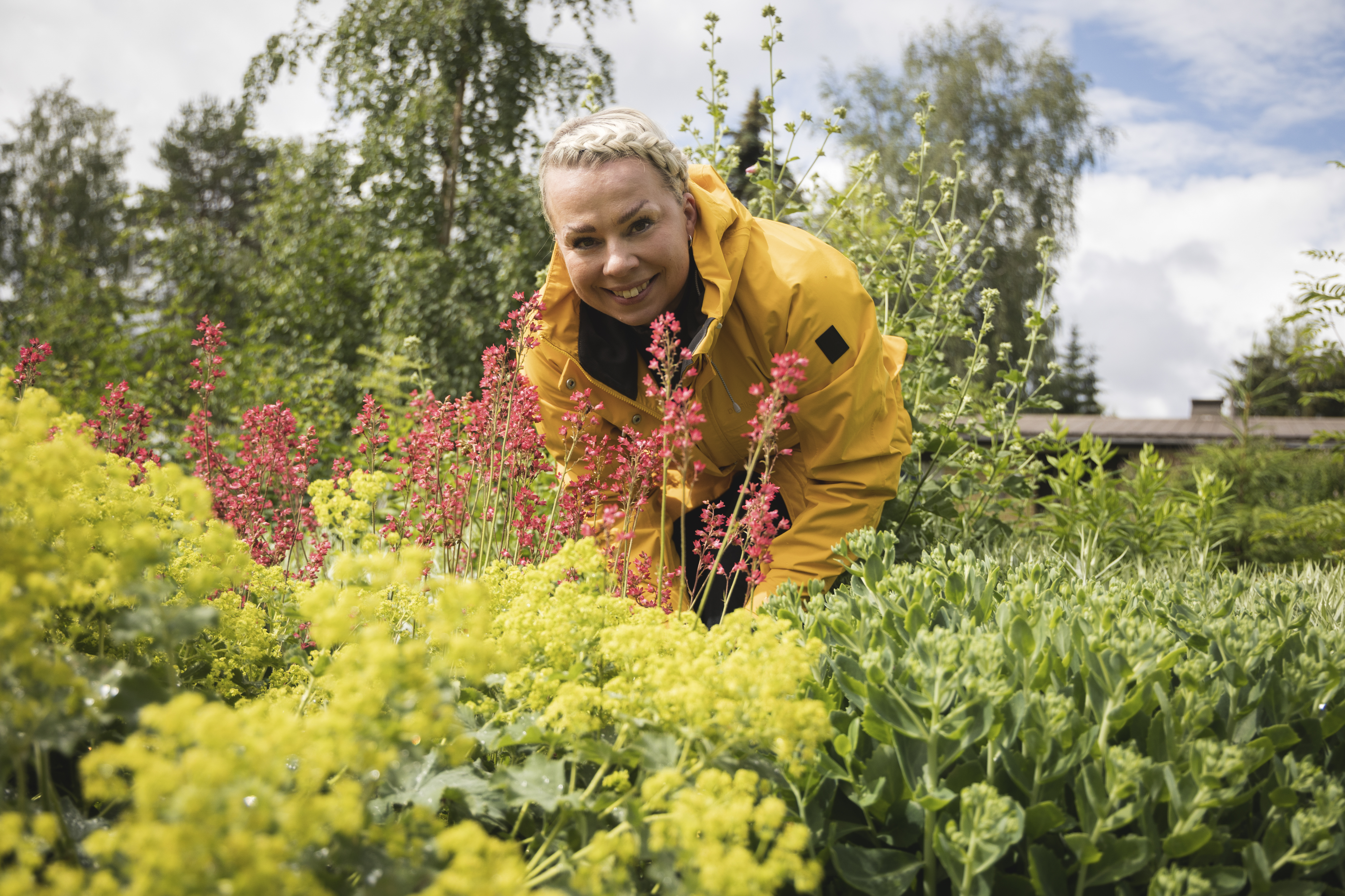 Keltatakkinen nainen hymyilee kauniin kukkapenkin takaa.