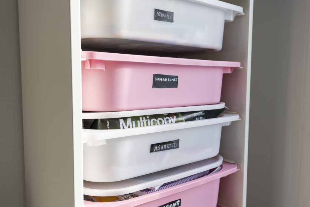 Valkoisia ja vaaleanpunaisia laatikoita kaapissa pystyrivissä. Laatikoissa lukee, mitä niissä on, mm. kyniä, hamahelmiä ja askartelutarvikkeita.