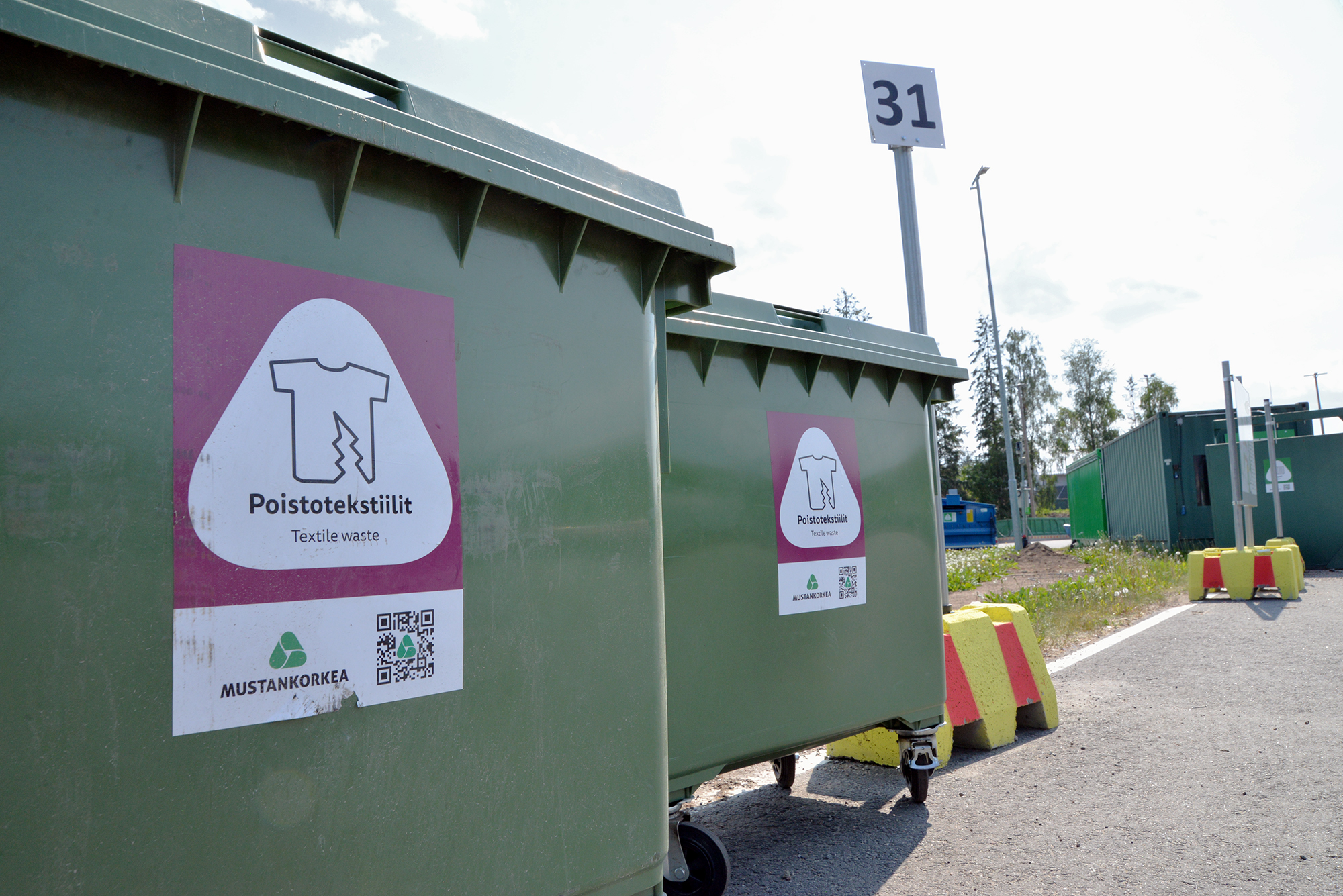 Kaksi vihreää jäteastiaa, joissa violetit poistotekstiilin jätetarrat. Yläpuolella lukee kierrätyspisteen numero 31.