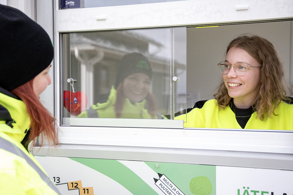 Kaksi naista katsoo toisiaan hymyillen. Kummallakin on huomiovaatteet, toinen istuu infokopin sisällä, toinen on pipo päässä ulkona.