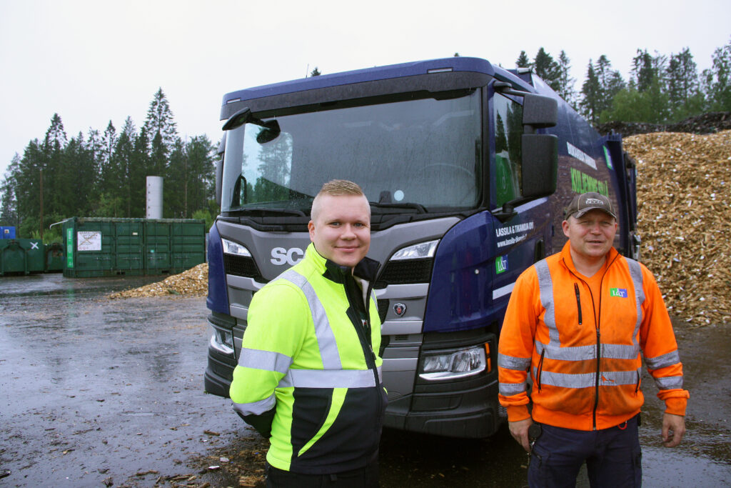 Miehet seisovat jäteauton edessä ja hymyilevät kameralle. Toisella on päällään keltainen takki, toisella oranssi. Ulkona on sateinen sää.