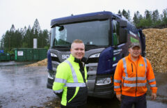 Miehet seisovat jäteauton edessä ja hymyilevät kameralle. Toisella on päällään keltainen takki, toisella oranssi. Ulkona on sateinen sää.