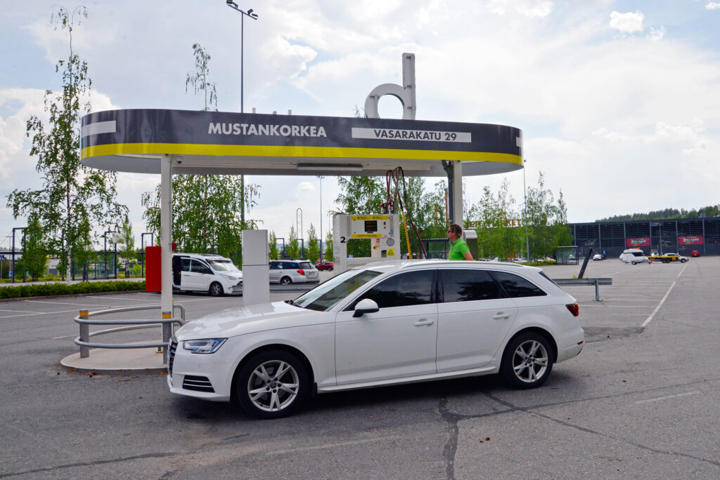 Valkoinen auto on tankkaamassa biokaasua. Taustalla näkyy Citymarketin liikekeskus.