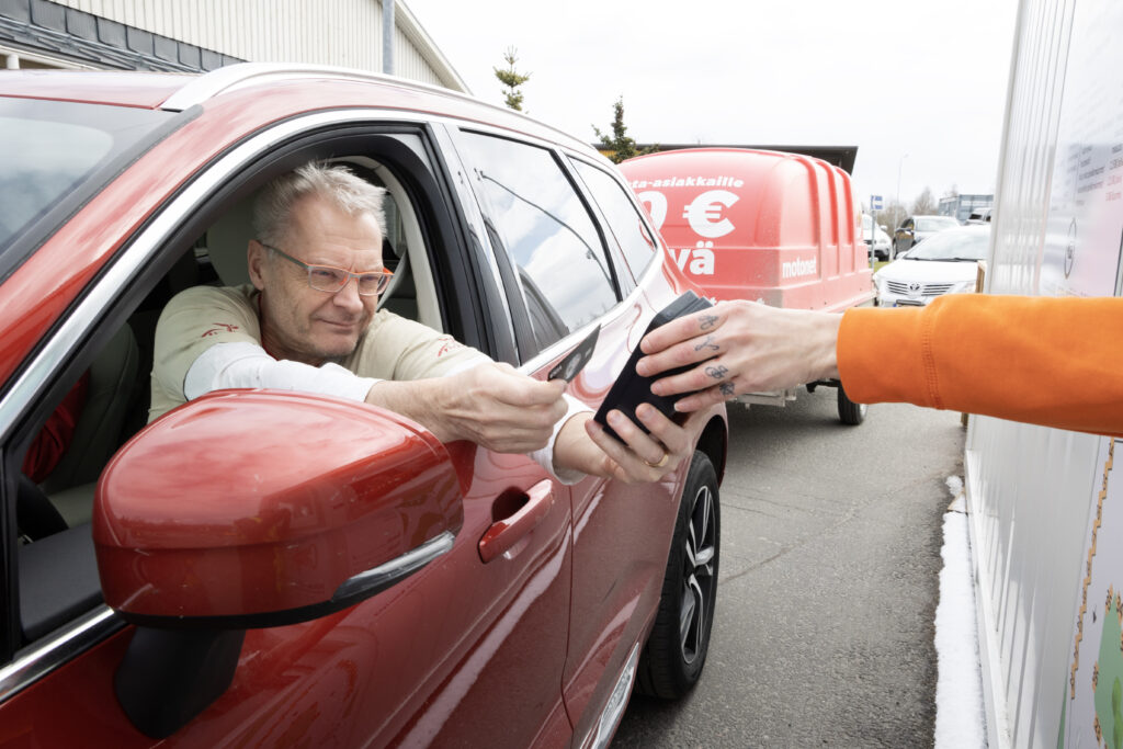 Punaisessa autossa oleva silmälasipäinen mies maksaa käynnistä kortilla, oikealta näkyy käsi, jossa maksuautomaatti.