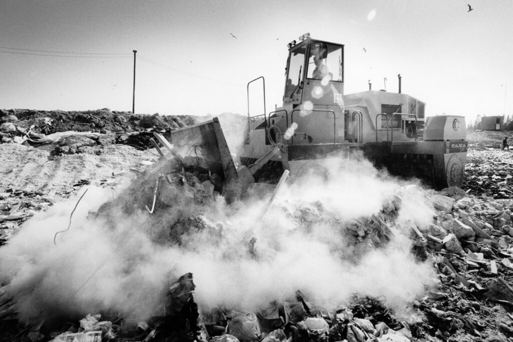 Mustavalkoisessa kuvassa Tanan kaatopaikkajyrä jyrää alleen jätteitä, jotka pöllyävät. Lokit lentävät taivaalla ja sähkölinja siintää horisontissa.