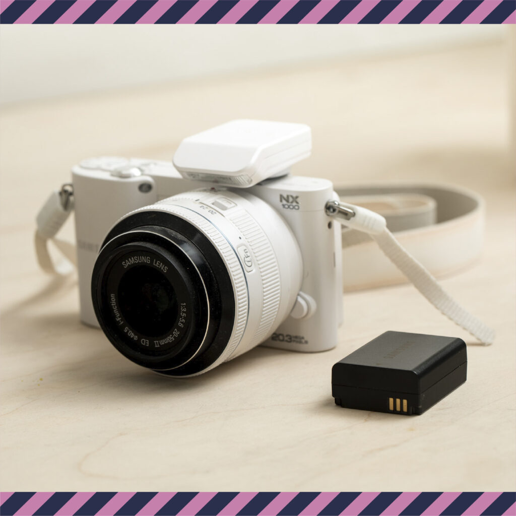 Valkoinen Samsungin kamera pöydällä, sen vieressä akku.