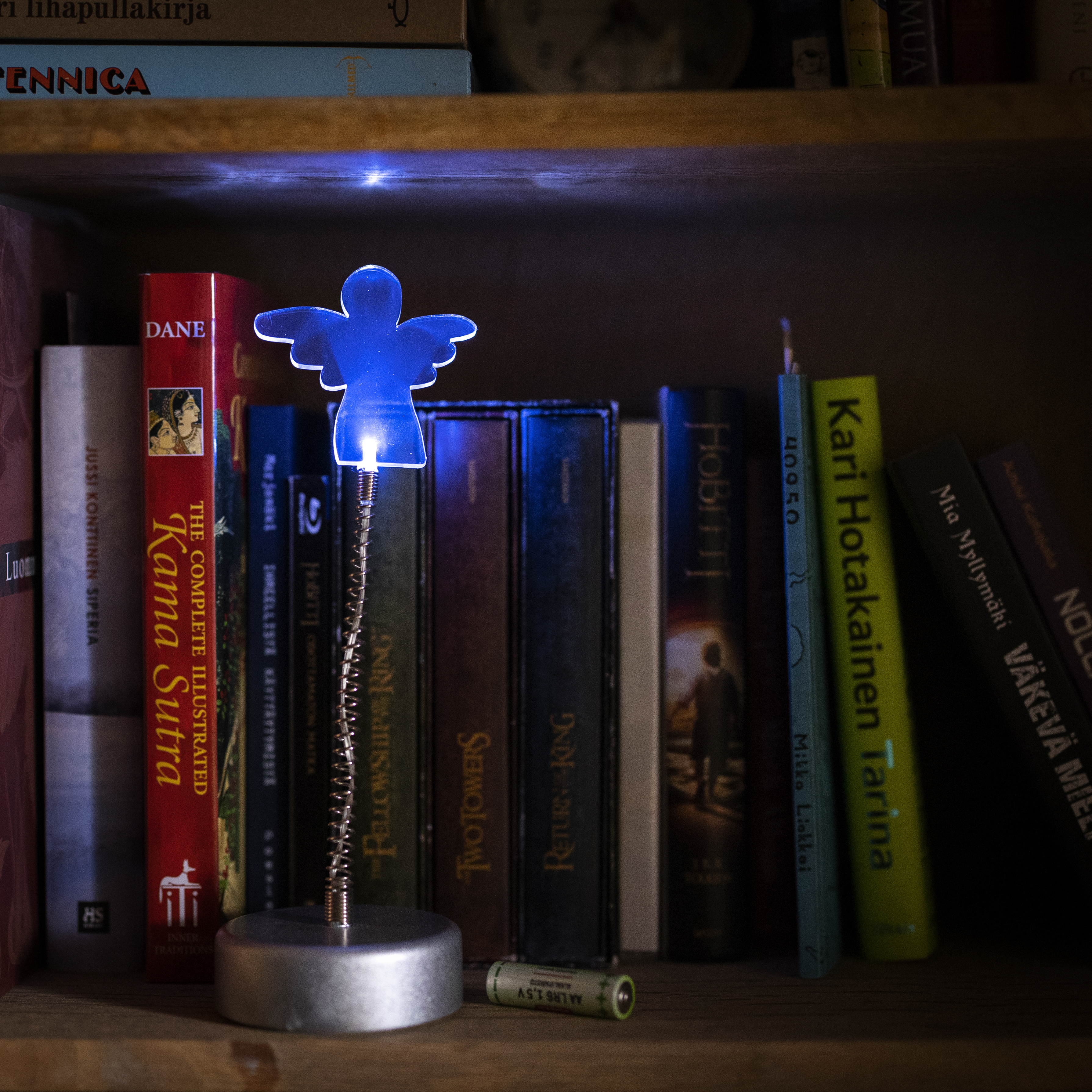 Enkelivalo loistaa sinisenä, takana näkyy kirjoja ja valon vieressä AA-paristo.