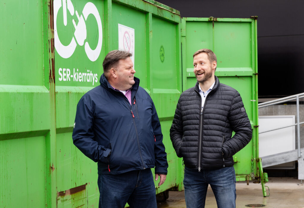 Kaksi miestä seisoo vihreän SER-kontin edessä ja katsoo toisiaan nauraen.