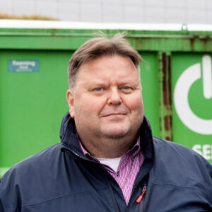 Mies katsoo kameraan ulkona. Taustalla näkyy vihreä jätekontti.