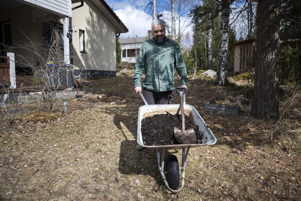 Mies työntää iloisena kottikärryjä, joissa on kyydissä multaa kompostorista. Taustalla näkyy omakotitaloa ja naapurin talo, on kevät.