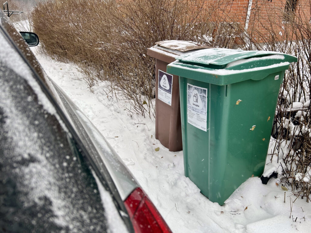 Vasemmalla näkyy osa harmaasta autosta ja oikealla kaksi jäteastiaa, sekajätteelle ja biojätteelle. Auton ja astian välissä on melko pieni tila.