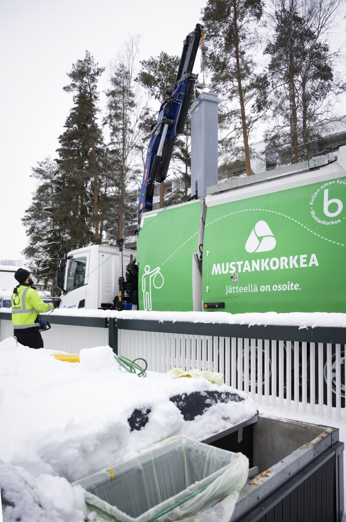 Mustankorkean jäteauto tyhjentää isoa syväkeräysastiaa. Etualalla näkyy roskiksia.