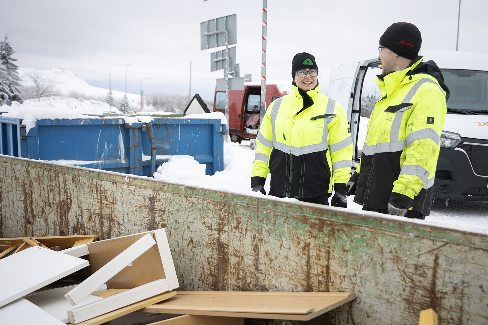 Nainen ja mies seisovat jätelavan edessä ja katsovat toisiaan hymyillen silmiin. Jätelavalla näkyy erilaista käsiteltyä puujätettä. On talvi ja lunta. Takana näkyy kaksi pakettiautoa.