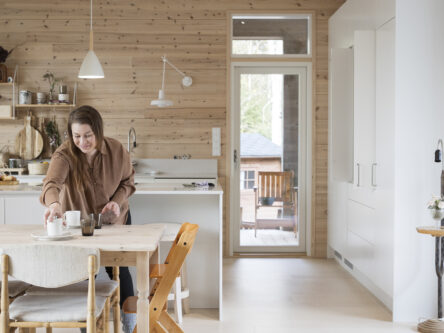 Nainen kattaa pöytään kahvikuppeja kotinsa keittiössä ja hymyilee.