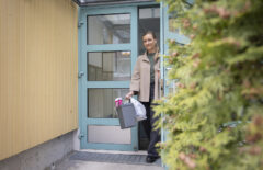 Nuori nainen avaa kerrostaloa ovea. Kädessään hänellä on sekajätteen roskapussi sekä ämpäri, jossa lehtiä ja muuta paperia. Etualalla näkyy taloyhtiön pihalla oleva havupuu.