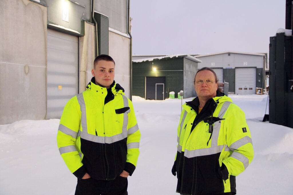 Nuori ja hieman vanhempi mies seisovat huomiovaatteet päällään talvisessa ympäristössä. Takana näkyy biokaasulaitos.