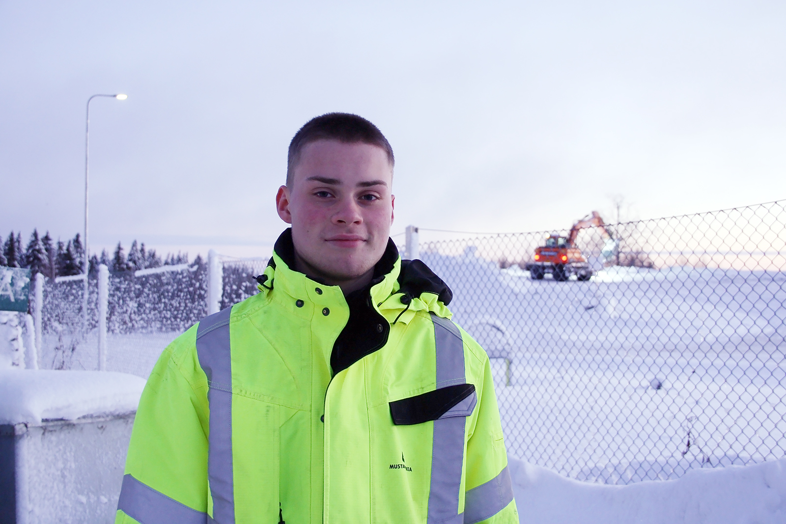 Nuori mies katso kameraan ja hymyilee. Hänellä on päällään keltainen huomiotakki. On kylmä talvi, taustalla töitä tekee kaivinkone.