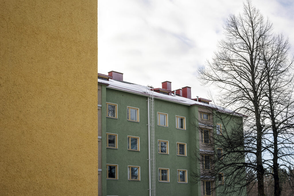 Edessä näkyy keltaisen rakennuksen seinää ja takana vihreä kerrostalo. Puissa ei ole enää lehtiä, taivas on pilvinen.