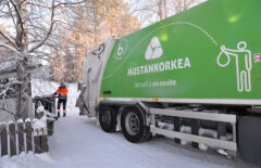 Etualalla on Mustankorkean vihreä jäteauto. Taaempana näkyy jäteauton kuljettaja, joka tuo jäteautolle isoa tummaa jäteastiaa. On kaunis ja aurinkoinen talvipäivä.