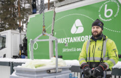 Mies pitää käsissään jäteauton nostorin ohjainta. Edessä näkyy iso syväkeräysastia, mitä nostetaan jäteastiasta. Takana näkyy vihreä Mustankorkean jäteauto, on talvi.