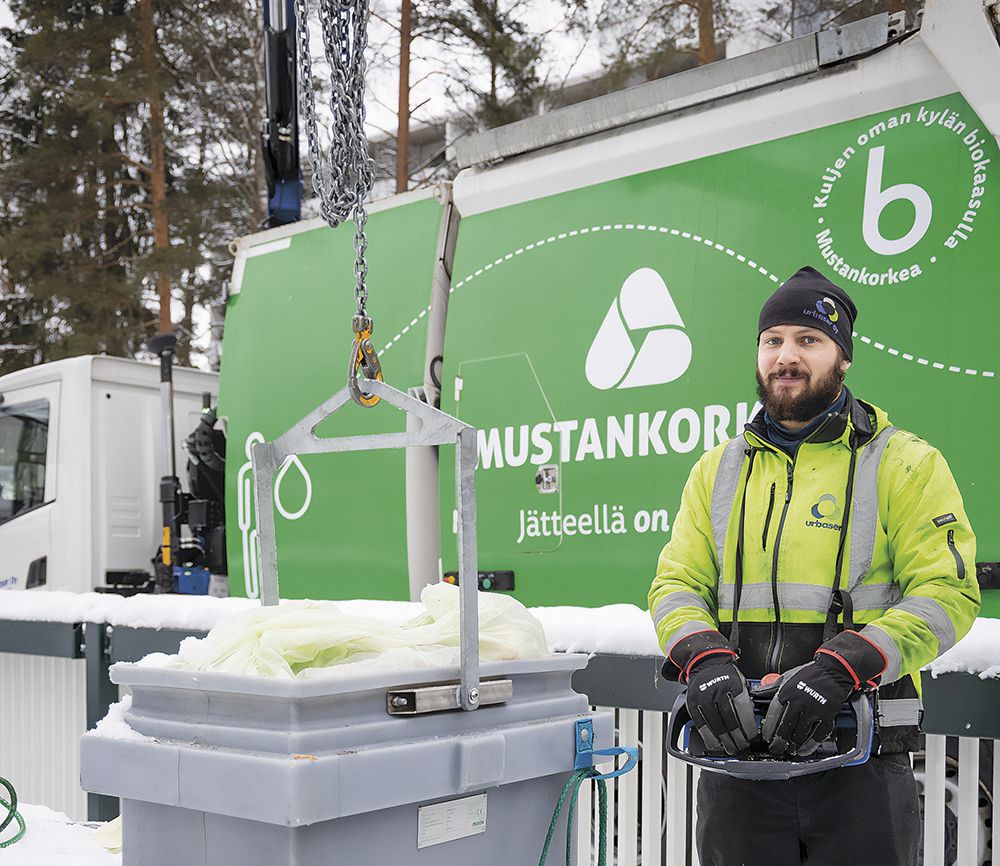 Mies pitää käsissään jäteauton nostorin ohjainta. Edessä näkyy iso syväkeräysastia, mitä nostetaan jäteastiasta. Takana näkyy vihreä Mustankorkean jäteauto, on talvi.