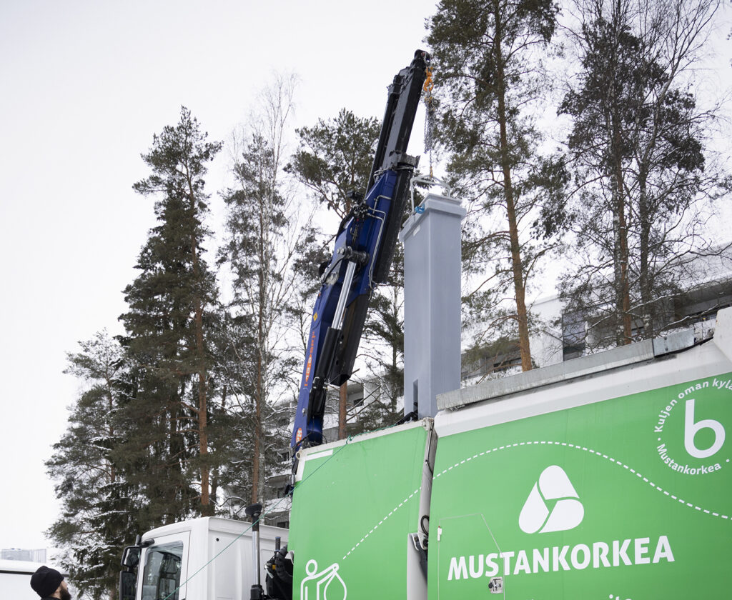 Kuvan alareunassa näkyy vihreää Mustankorkean jäteautoa. Kuvan keskellä on nostori, josta roikkuu iso harmaa jäteastia.