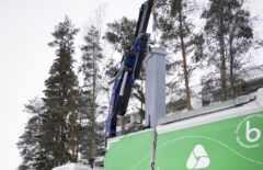 Kuvan alareunassa näkyy vihreää Mustankorkean jäteautoa. Kuvan keskellä on nostori, josta roikkuu iso harmaa jäteastia.