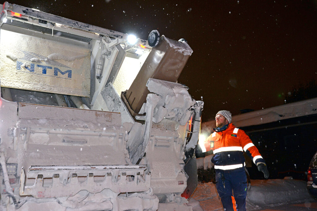 Lunta sataa, kun jäteauto tyhjentää pimeässä jäteastiaa. Kuljettajalla on suojalasit.