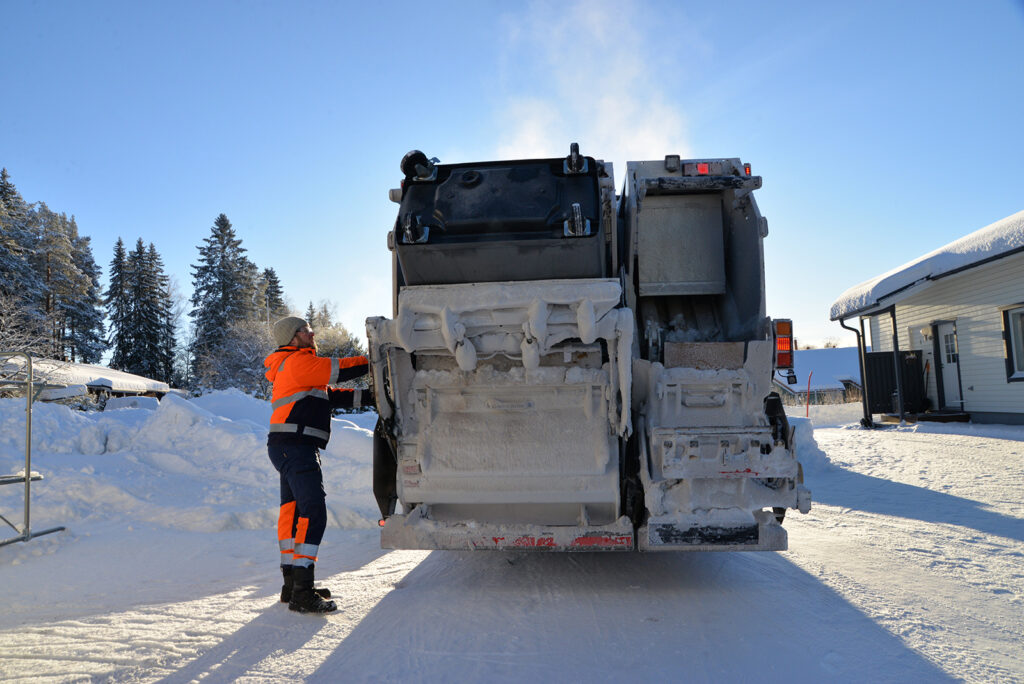 Oransseihin huomiovaatteisiin pukeutunut mies seisoo jäteauton vieressä. Jäteautossa on iso jäteastia, jota tyhjennetään autoon.