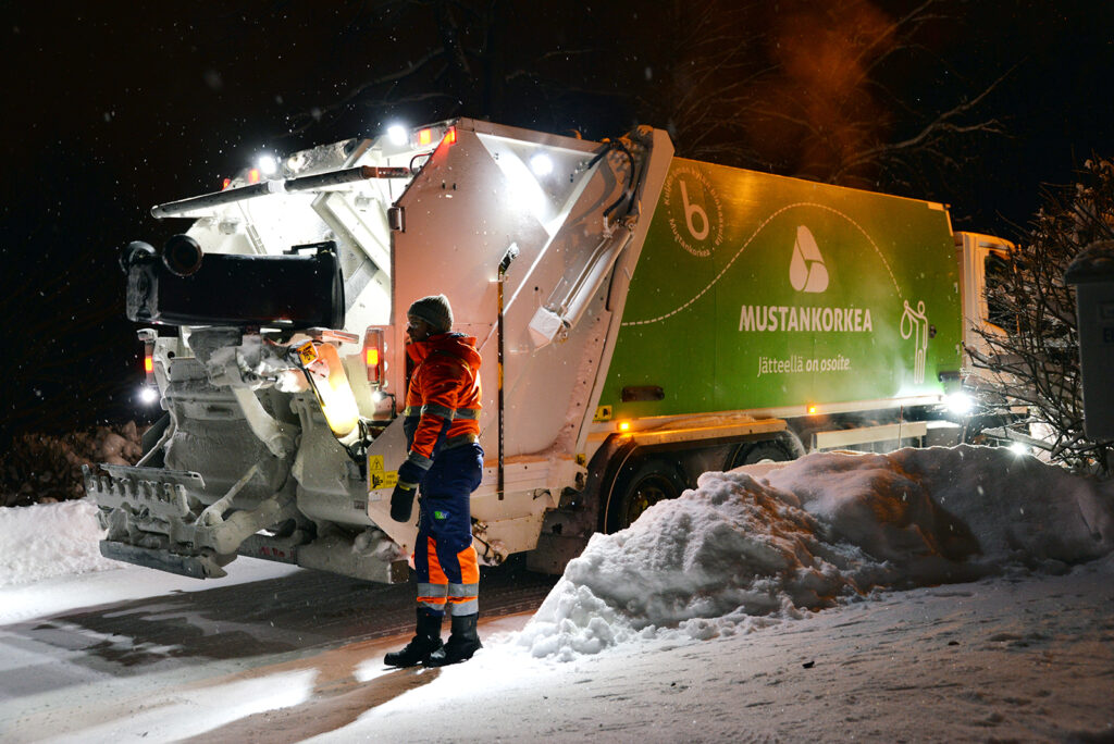 Mies tyhjentää jäteastiaa. On hyvin pimeää. Jäteauton valot valaisevat jäteauton takaosaa.