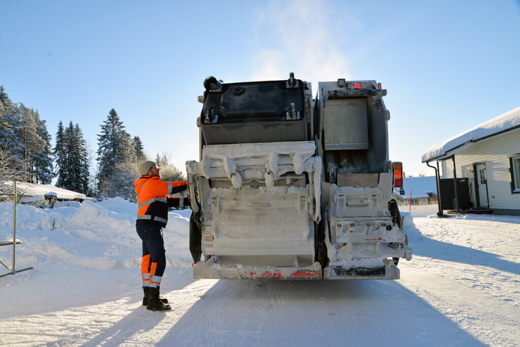 Jäteauton kuljettaja tyhjentää isoa mustaa jäteastiaa. On kaunis aurinkoinen pakkaspäivä.