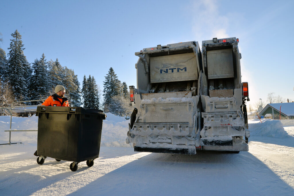 Jäteauton kuljettaja vie isoa jäteastiaa jäteautolle. On kaunis talvipäivä.
