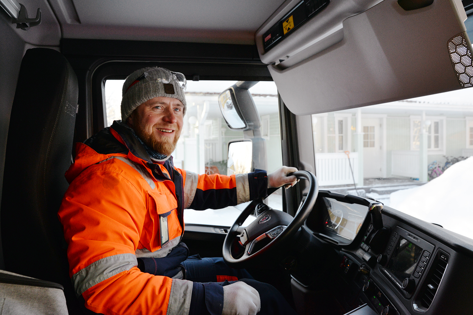 Huomiovaatteissa oleva jäteauton kuljettaja hymyilee ja katsoo ulos ikkunasta.