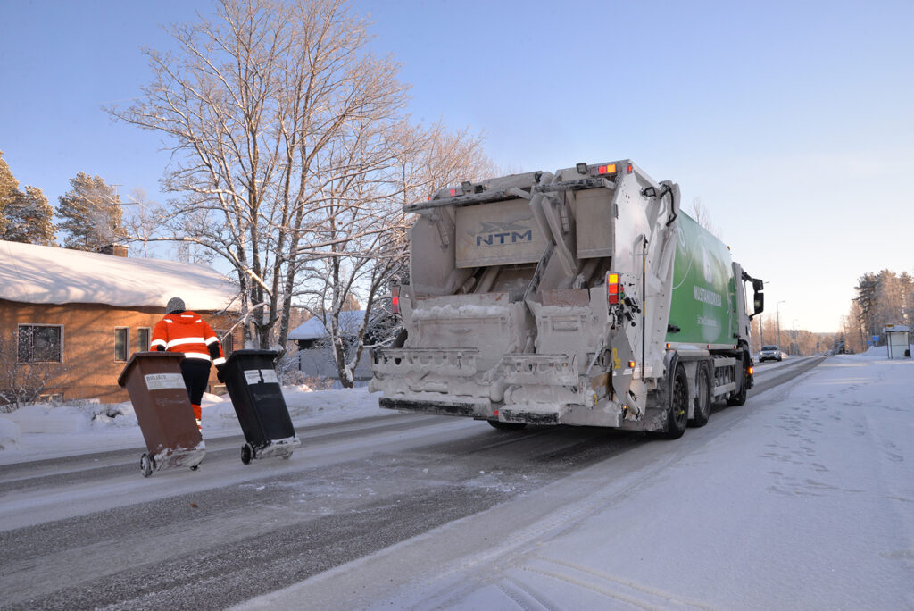 Mies vetää perässään roskiksia autotien yli. Jäteauton takaa tulee henkilöauto, on talvi.